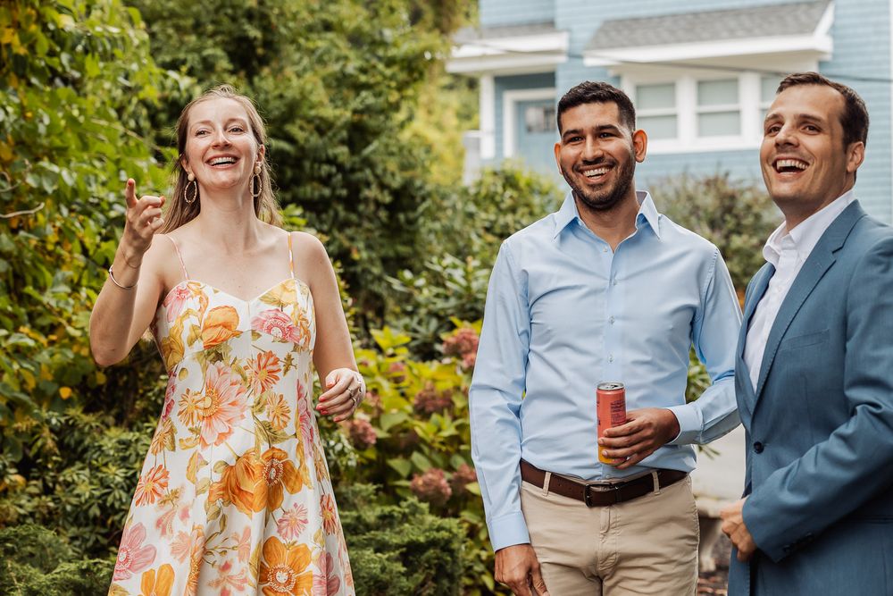 Three people share laughter during an outdoor gathering near a blue house.