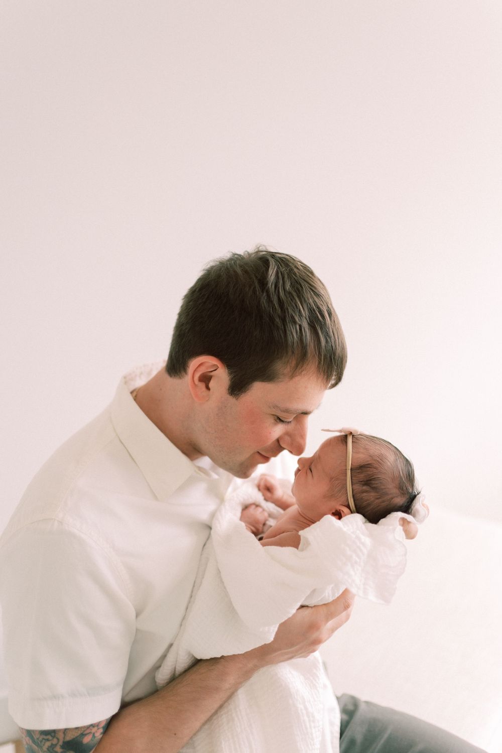 A tender moment between a parent and newborn baby dressed in white against a light background.