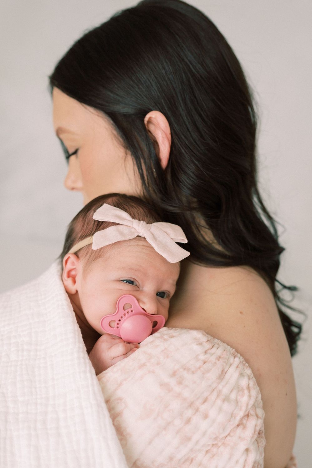 A sleeping baby wearing a pink bow headband and pacifier rests against a parent in soft pink tones.