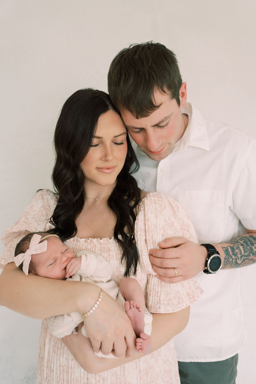 A couple in white and pink holds their newborn baby in a tender family portrait against a neutral background.