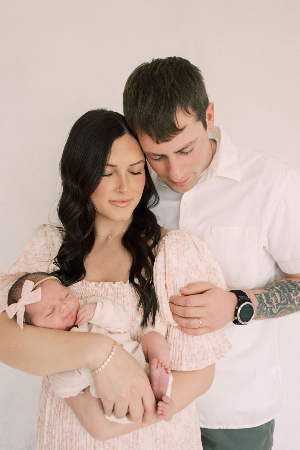 A tender family moment as parents in light colors cradle their newborn baby against a neutral background.