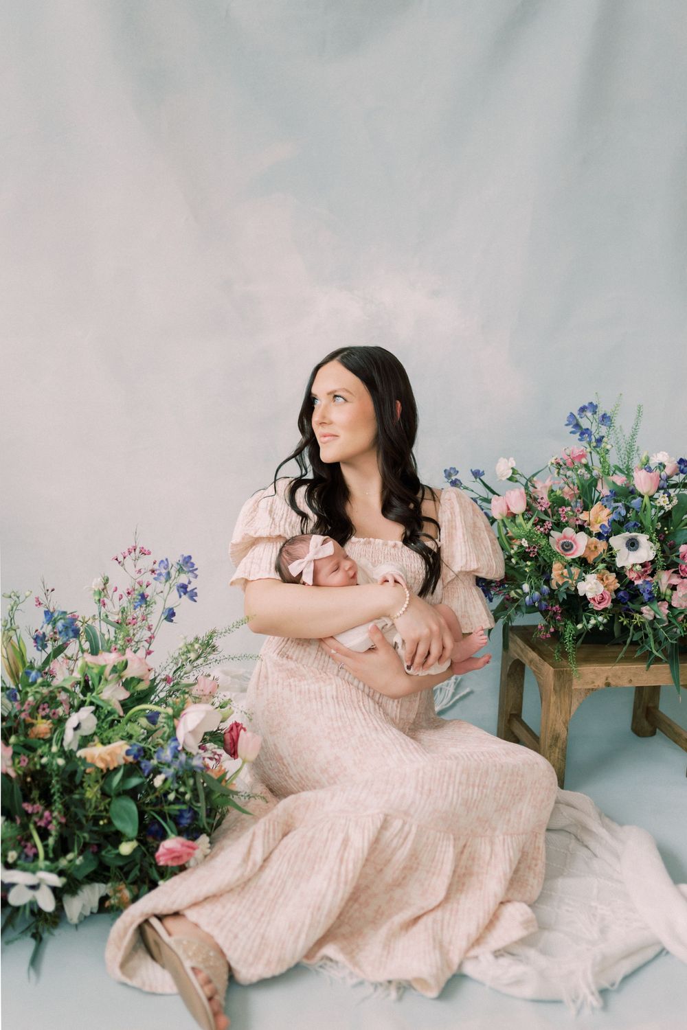 Woman in pink gown poses with infant amid floral arrangements against light backdrop.