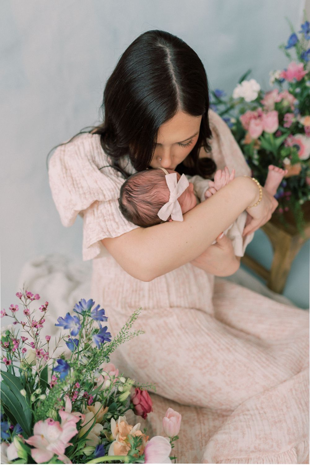 A woman in a pink dress holds a baby surrounded by colorful spring flowers in a dreamy studio portrait setting.