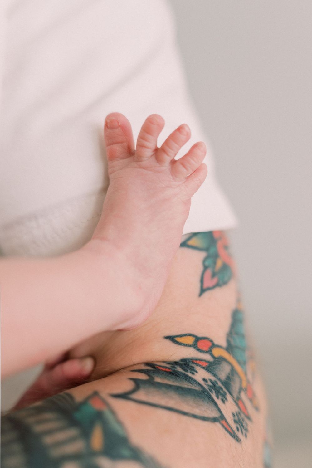 Baby's tiny hand rests against a tattooed arm showing colorful floral and nature-inspired designs.