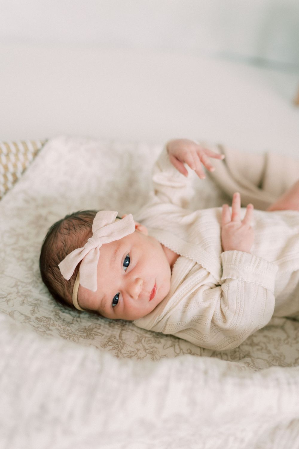 A newborn baby wearing a cream sweater and bow headband lies on soft white bedding.
