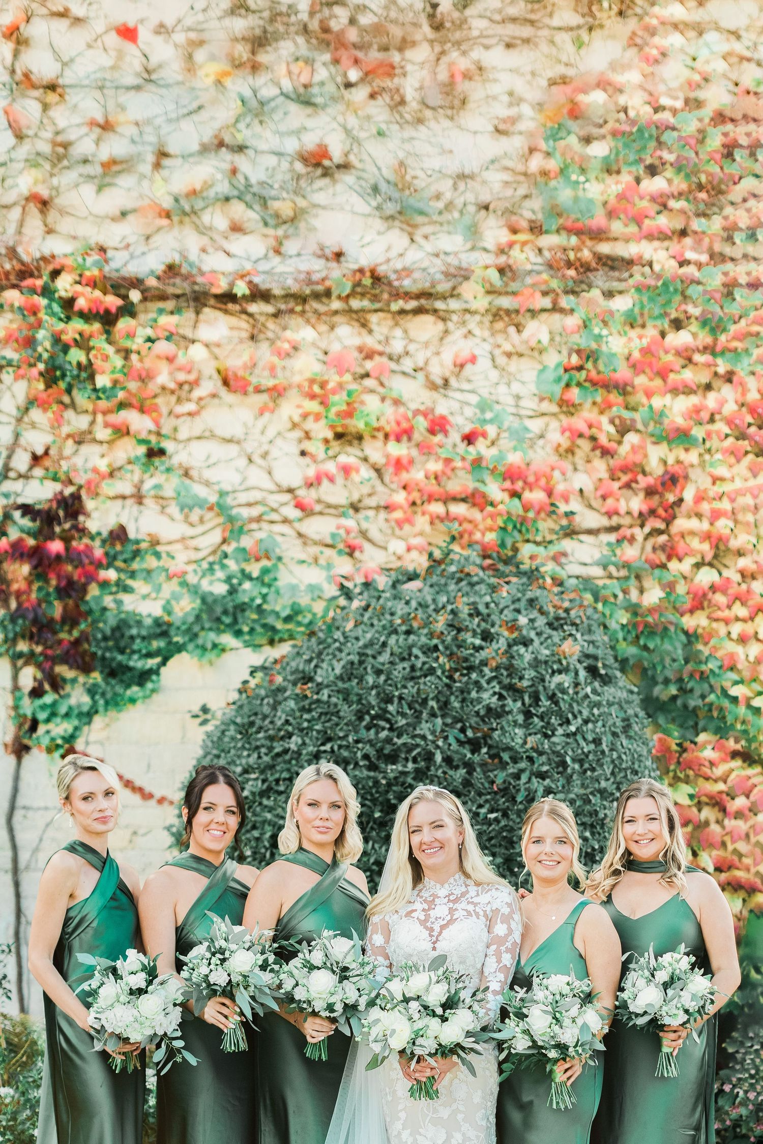 Wedding party in emerald green dresses and white bouquets against a wall covered in coral vines. Ellenborough Park by Hayley Elizabeth