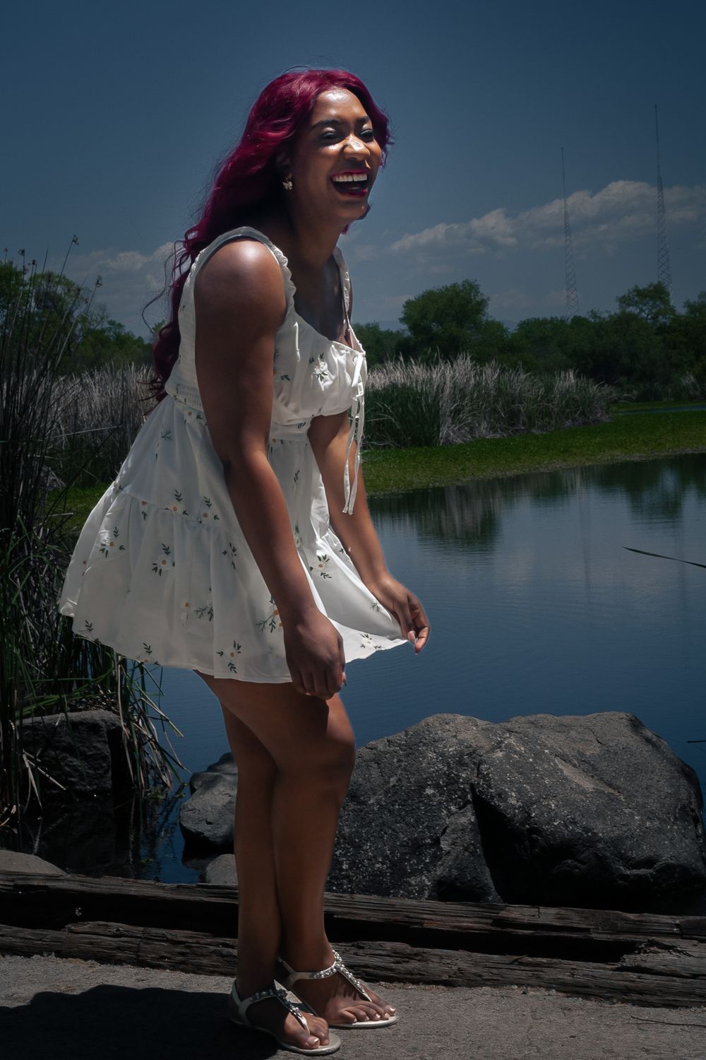 A person in a white sundress laughs joyfully while standing near a lake on a sunny summer day.