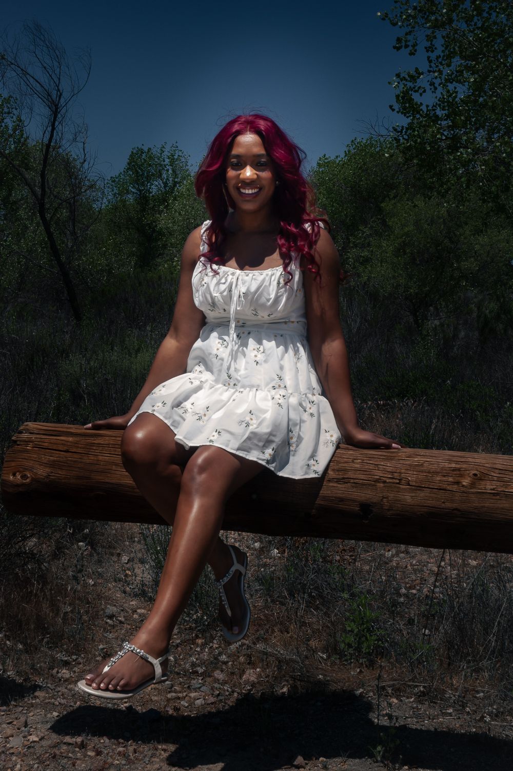 Someone with vibrant magenta hair wearing a white summer dress sits on a wooden log in a wooded outdoor setting.