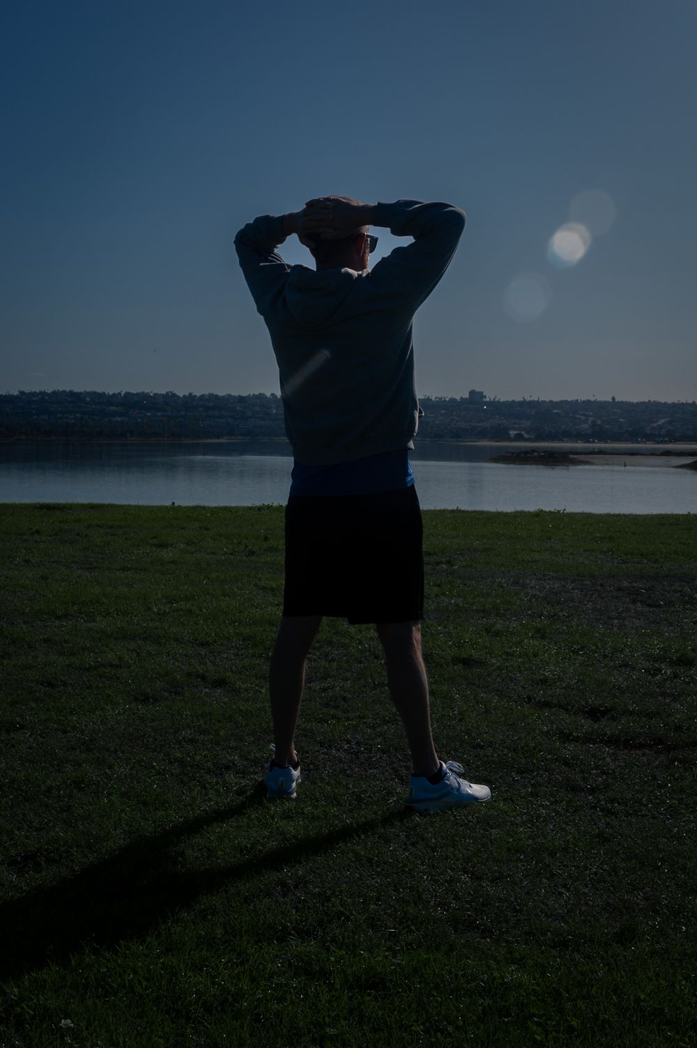Silhouetted figure stretches arms above head while standing on grassy hill overlooking water. Lifestyle photography, San Diego, California