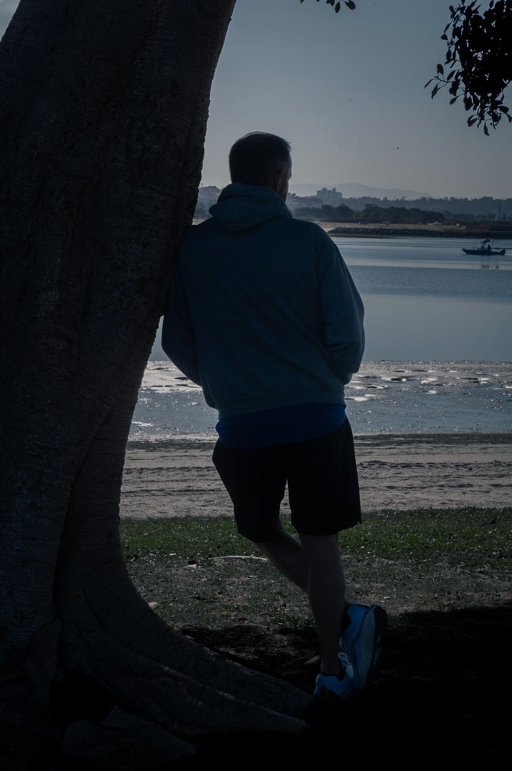 Person in blue jacket stands by tree silhouetted against waterfront view. Lifestyle photography, San Diego, California