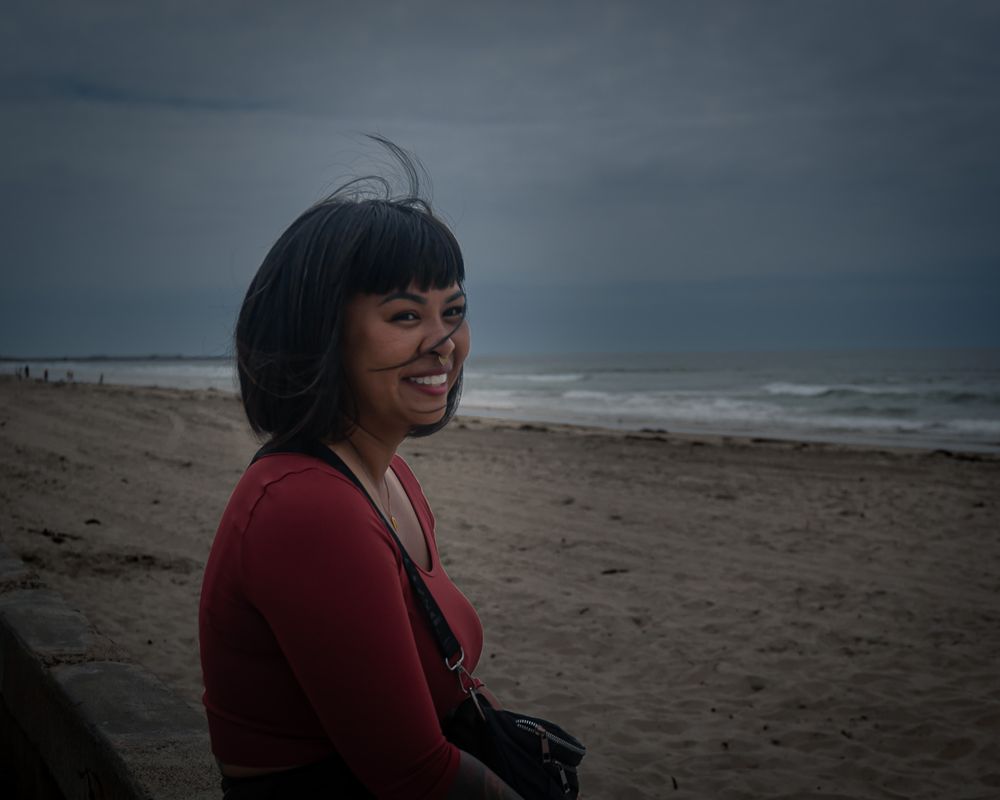 Someone in a red sweater smiles on a cloudy beach with stormy waves in the background.