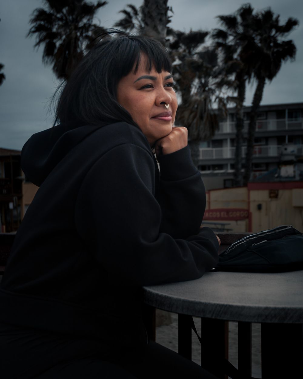 A person in a black hoodie smiles while resting at an outdoor table with palm trees in the evening.