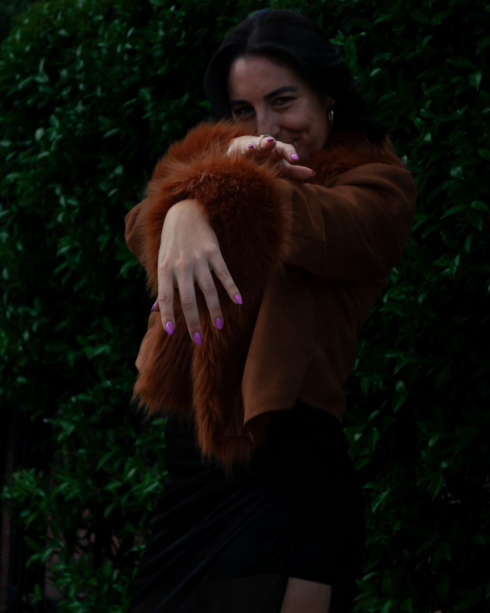 Person in brown fuzzy coat poses playfully with pink nail polish against dark green background. Lifestyle photography, San Diego, California