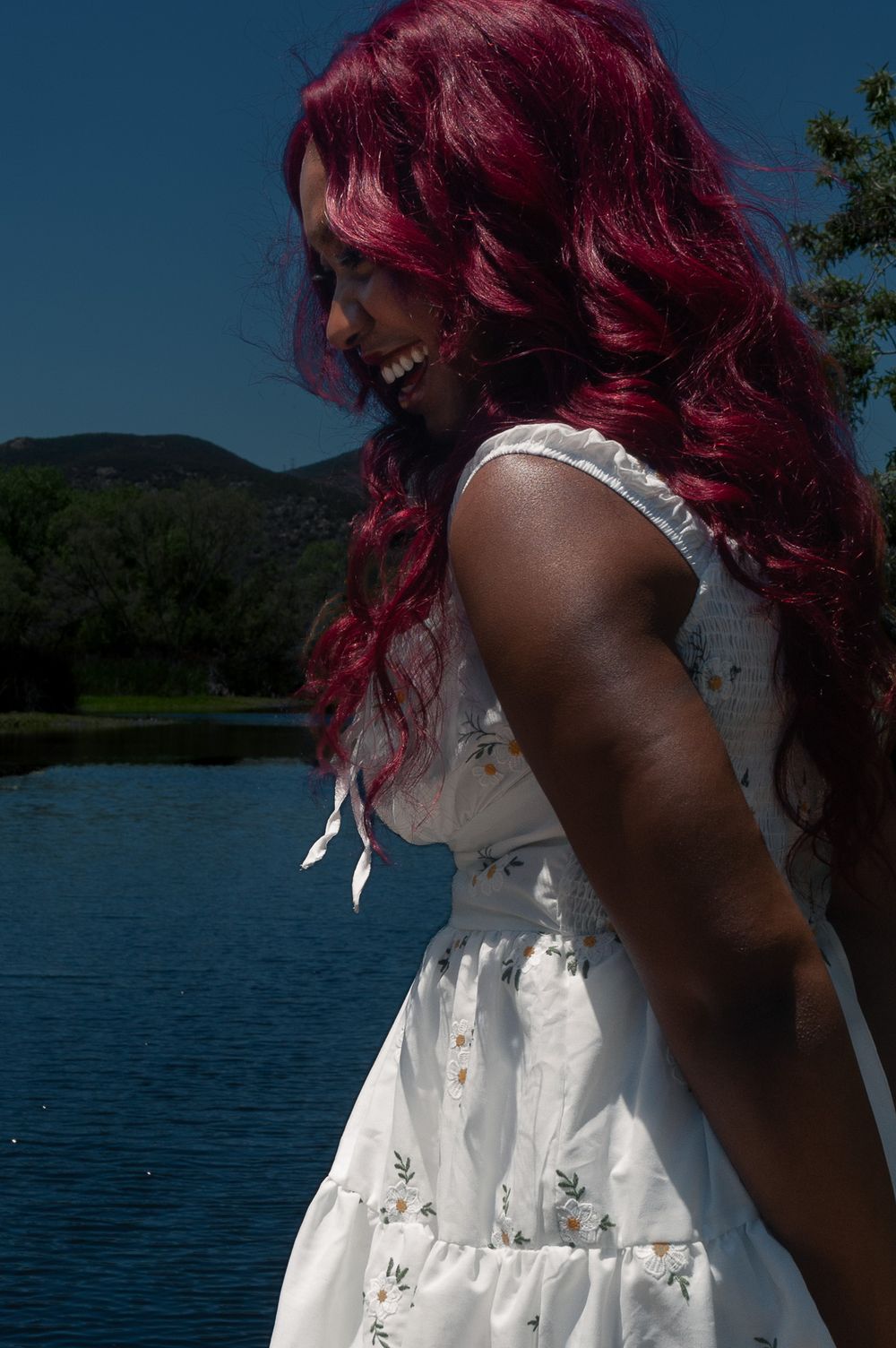 Person with vibrant red hair wearing a white sundress laughs while standing by a serene lake on a sunny day.