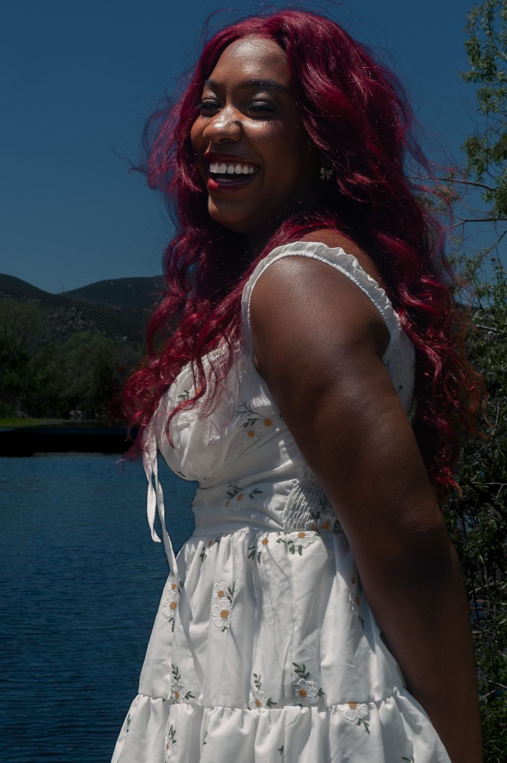 A joyful person with vibrant magenta hair wearing a white floral sundress stands near a serene lake on a sunny day.