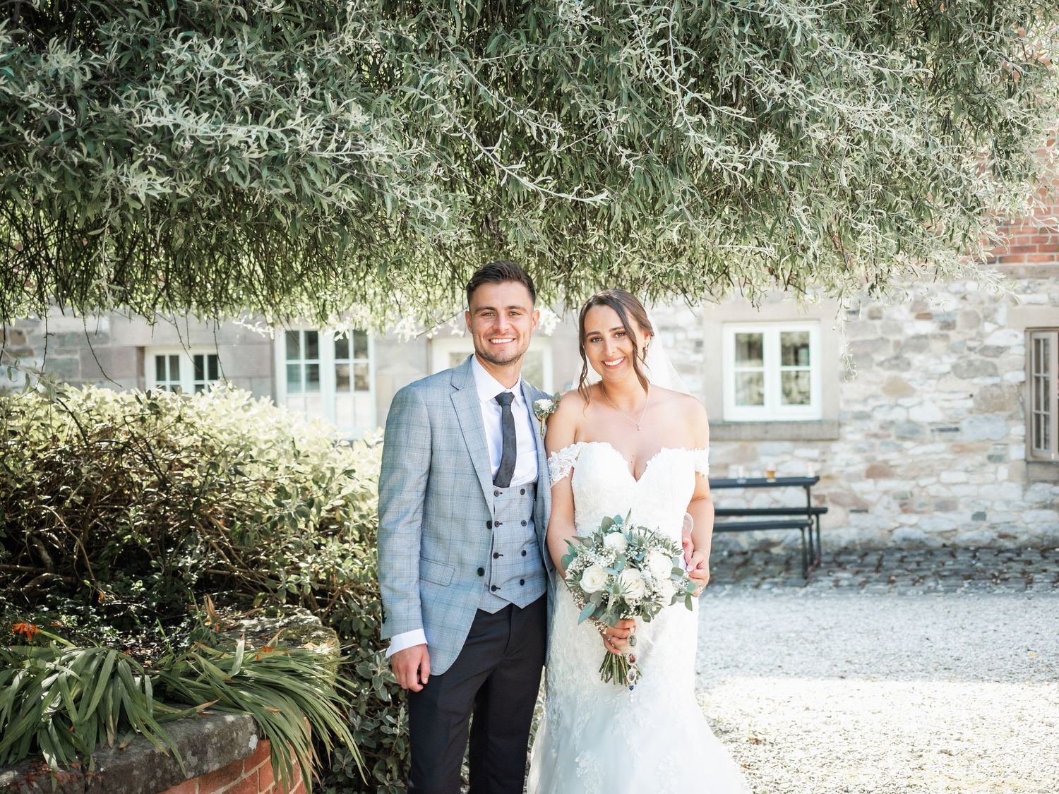 A bride and groom stand together at a rustic stone venue with overhanging foliage and white windows behind them.