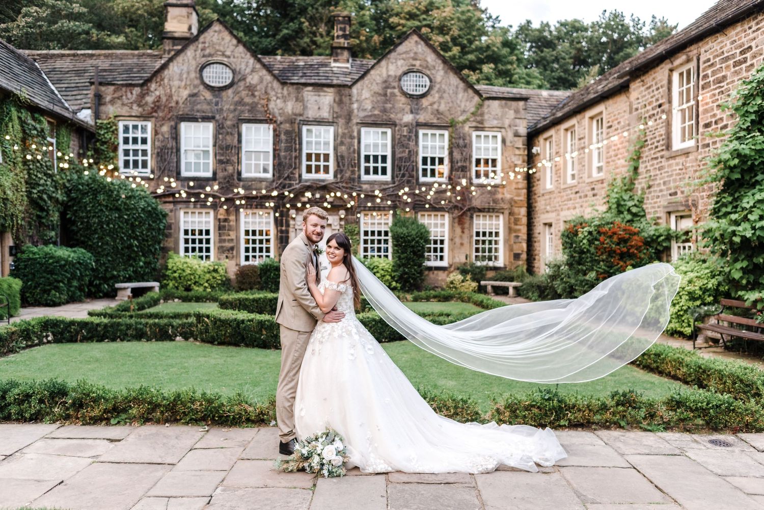 Elegant couple stands in front of historic stone mansion with beautiful landscaped gardens as veil flows in the wind.