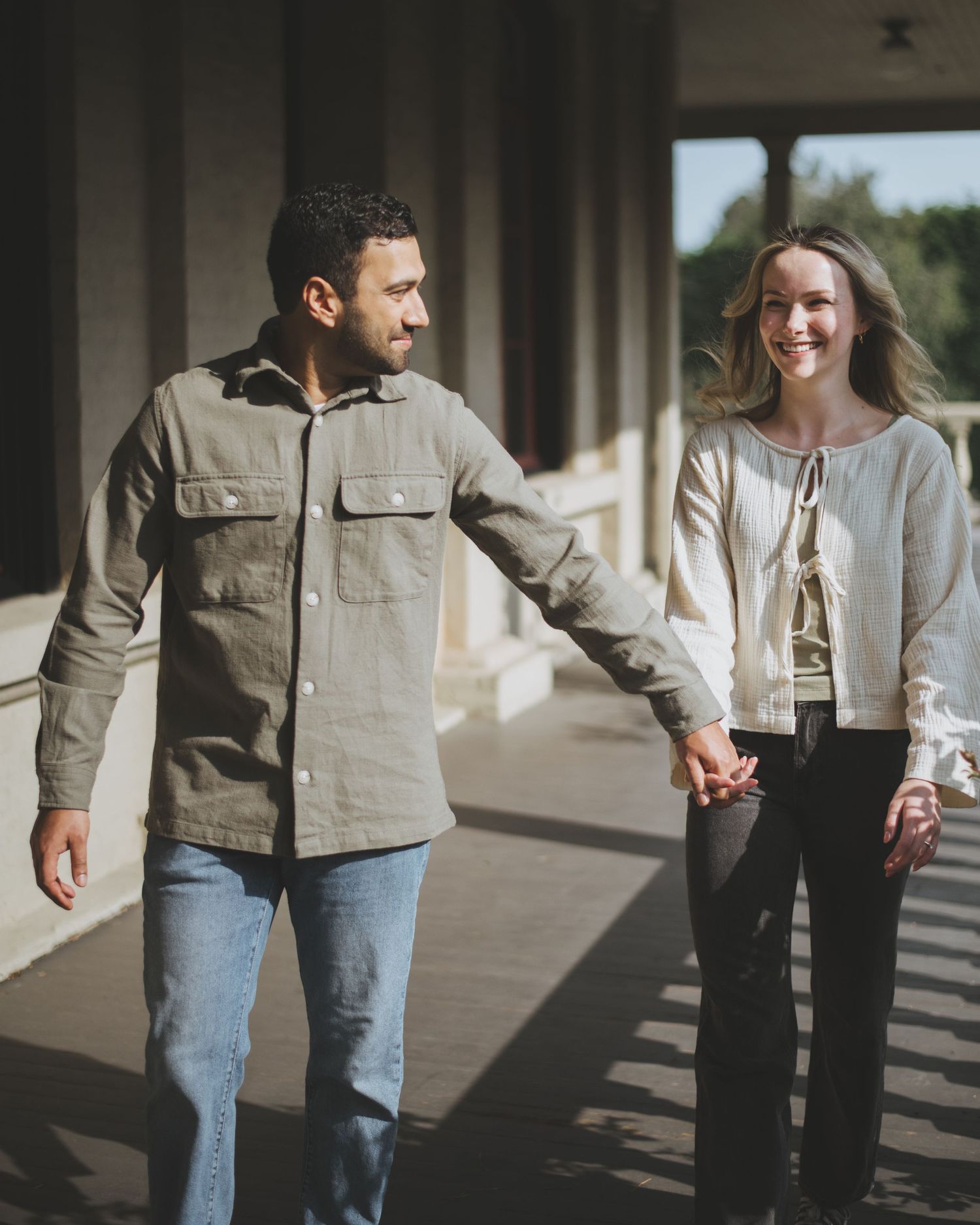 A couple walks hand in hand along a covered porch with columns and balustrades on a sunny day.