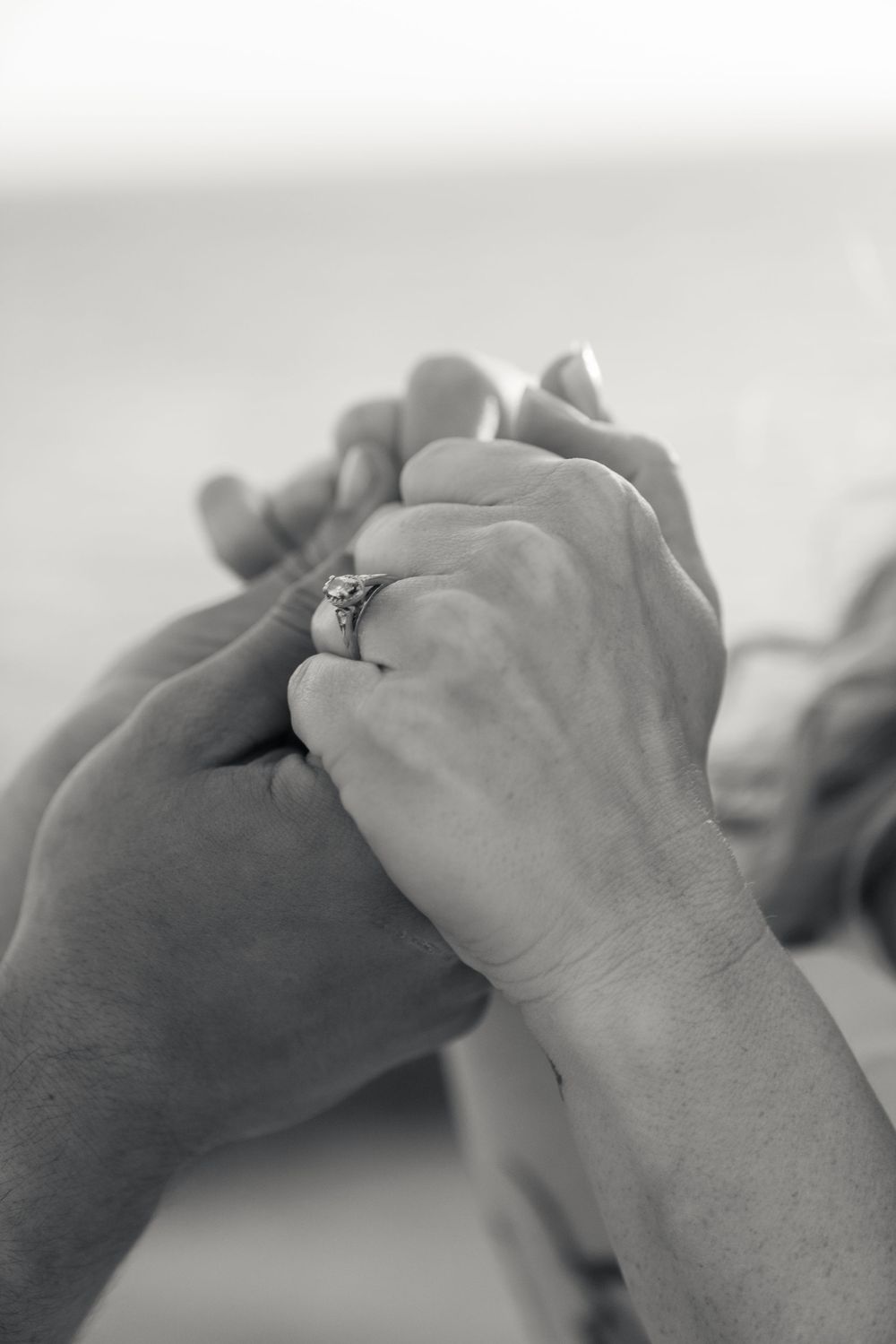 A close-up black and white photo of clasped hands with a diamond engagement ring.