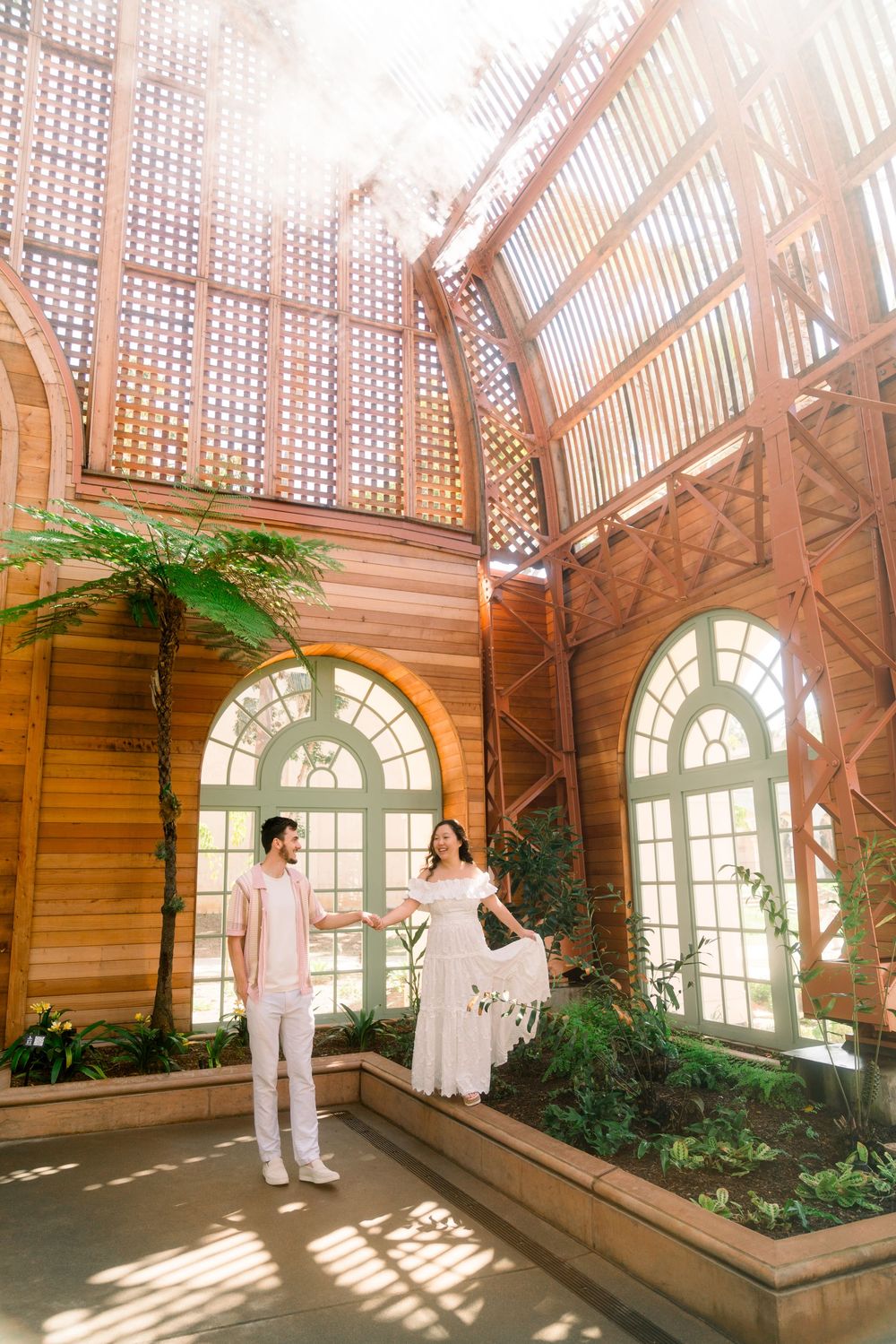 Two people in light outfits stand in a sunlit conservatory with arched windows and wooden lattice walls surrounded by plants.