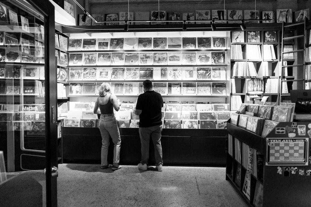 Two silhouetted customers browse a well-lit magazine and newspaper stand display in a black and white photograph.