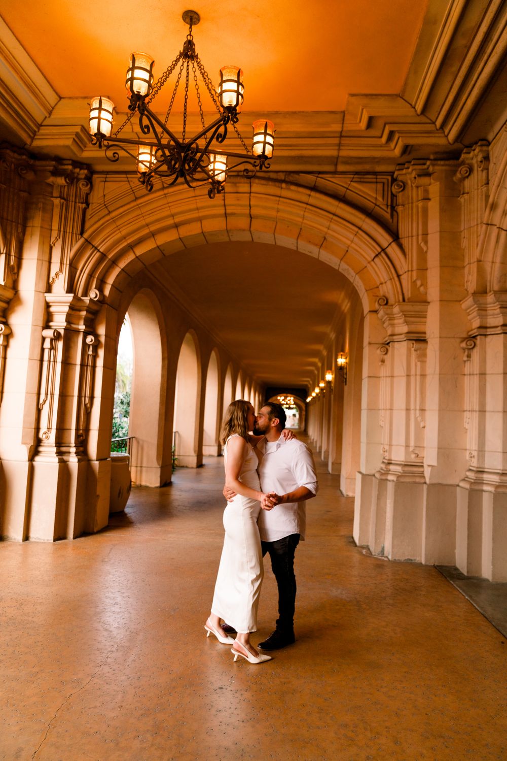A couple embraces in an elegant arched hallway with warm lighting and ornate architectural details.