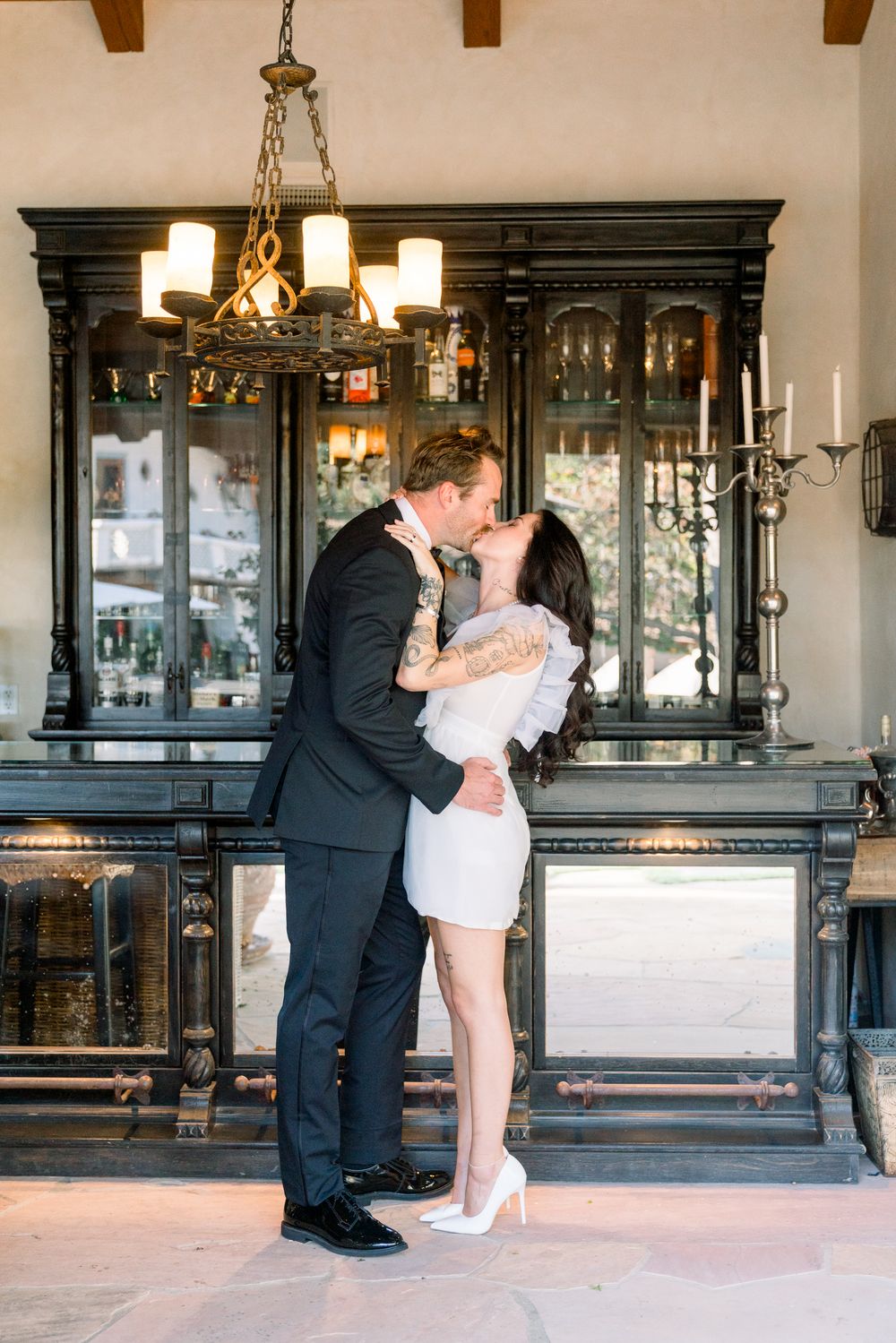 A romantic couple embraces in an elegant room with dark wooden furniture and chandelier lighting.