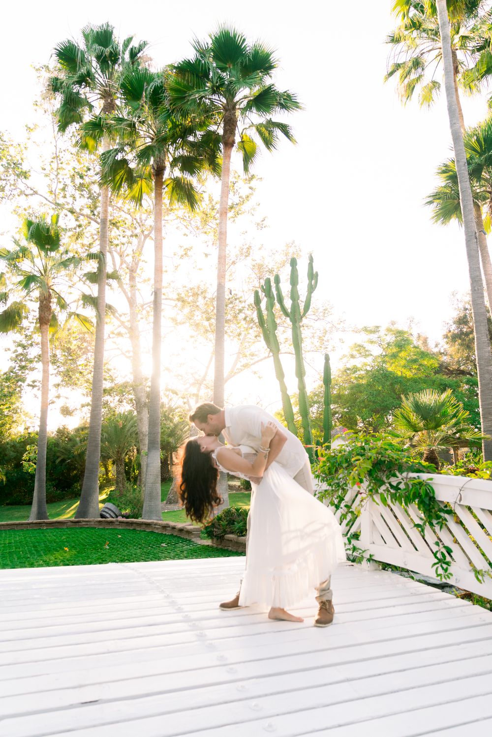 Couple shares romantic dip kiss pose on white deck surrounded by tropical palm trees and desert cacti at sunset.