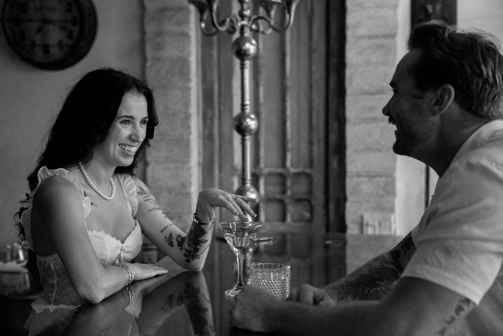 A black and white candid photo shows a couple sharing a conversation at a table in an elegant setting with ornate columns.