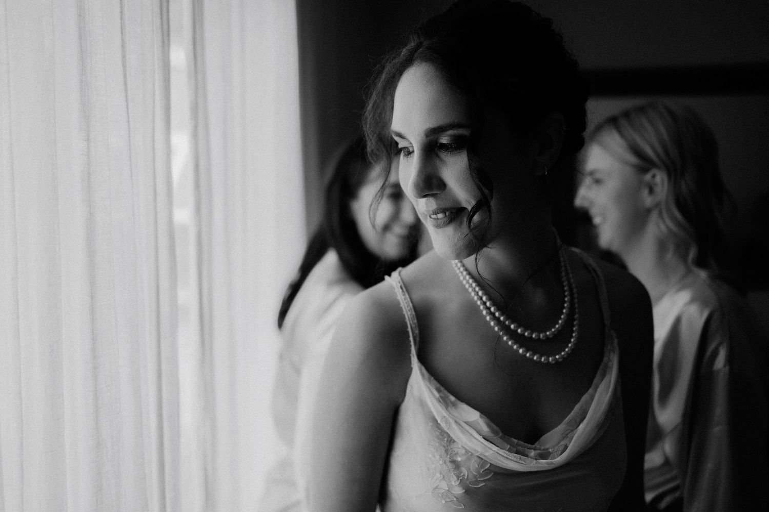 Black and white candid moment shows a bride wearing pearls smiling by a window with others in background.