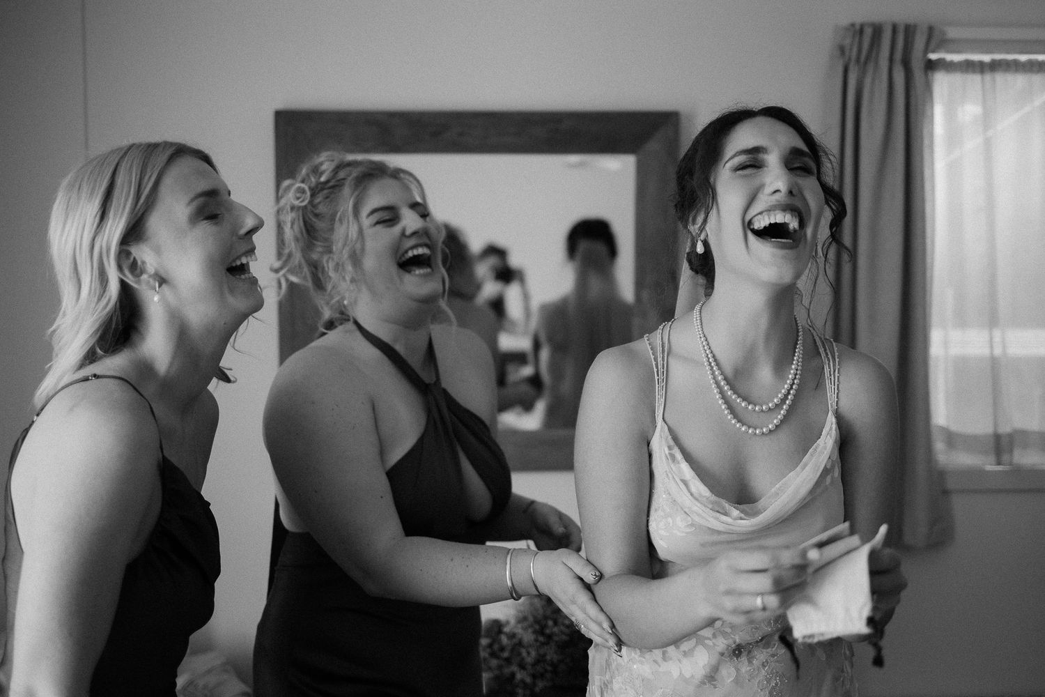 A candid black and white photo captures three women laughing together during a joyous moment at an indoor gathering.
