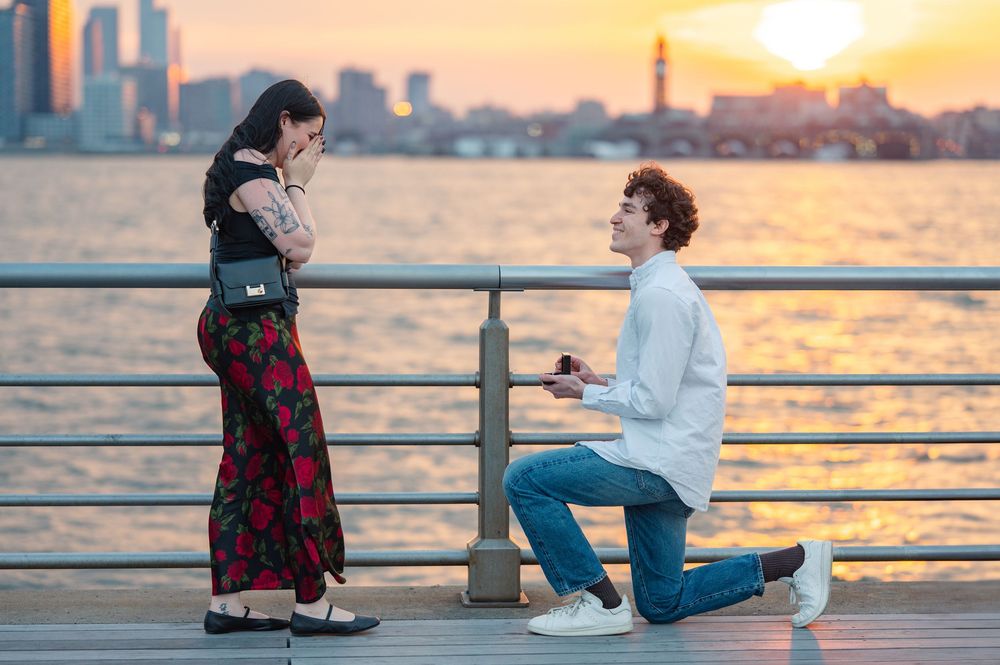 A surprise proposal in NYC silhouetted against a golden sunset on a waterfront pier with city skyline in the background.