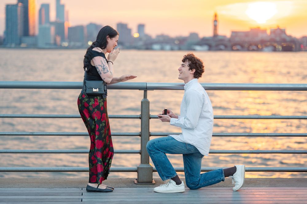A romantic proposal scene at sunset on a pier with the city skyline silhouetted in the background on Pier 45 at Hudson River Park in nYC