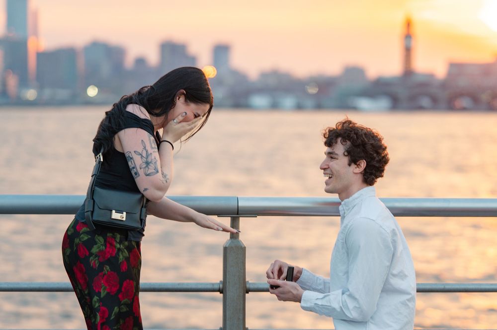 A romantic proposal scene on a waterfront pier at sunset with the city skyline glowing in the background in New York City