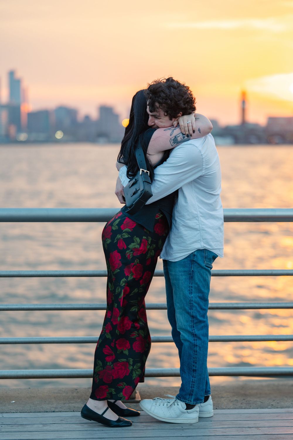 Couple embraces at sunset on waterfront pier with city skyline and orange sky in background after their surprise proposal