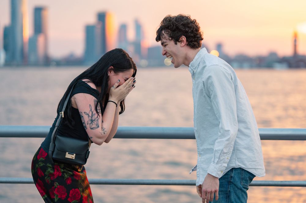 Two people share an emotional moment at sunset on a waterfront pier with a cityscape in the background after their proposal in NYC