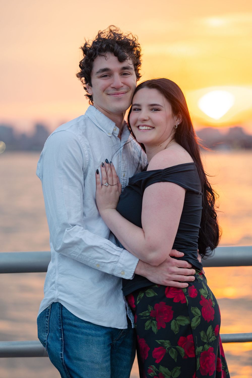 A newly engaged couple embracing at sunset on a pier with water and cityscape behind them at Pier 45 at Hudson River Park