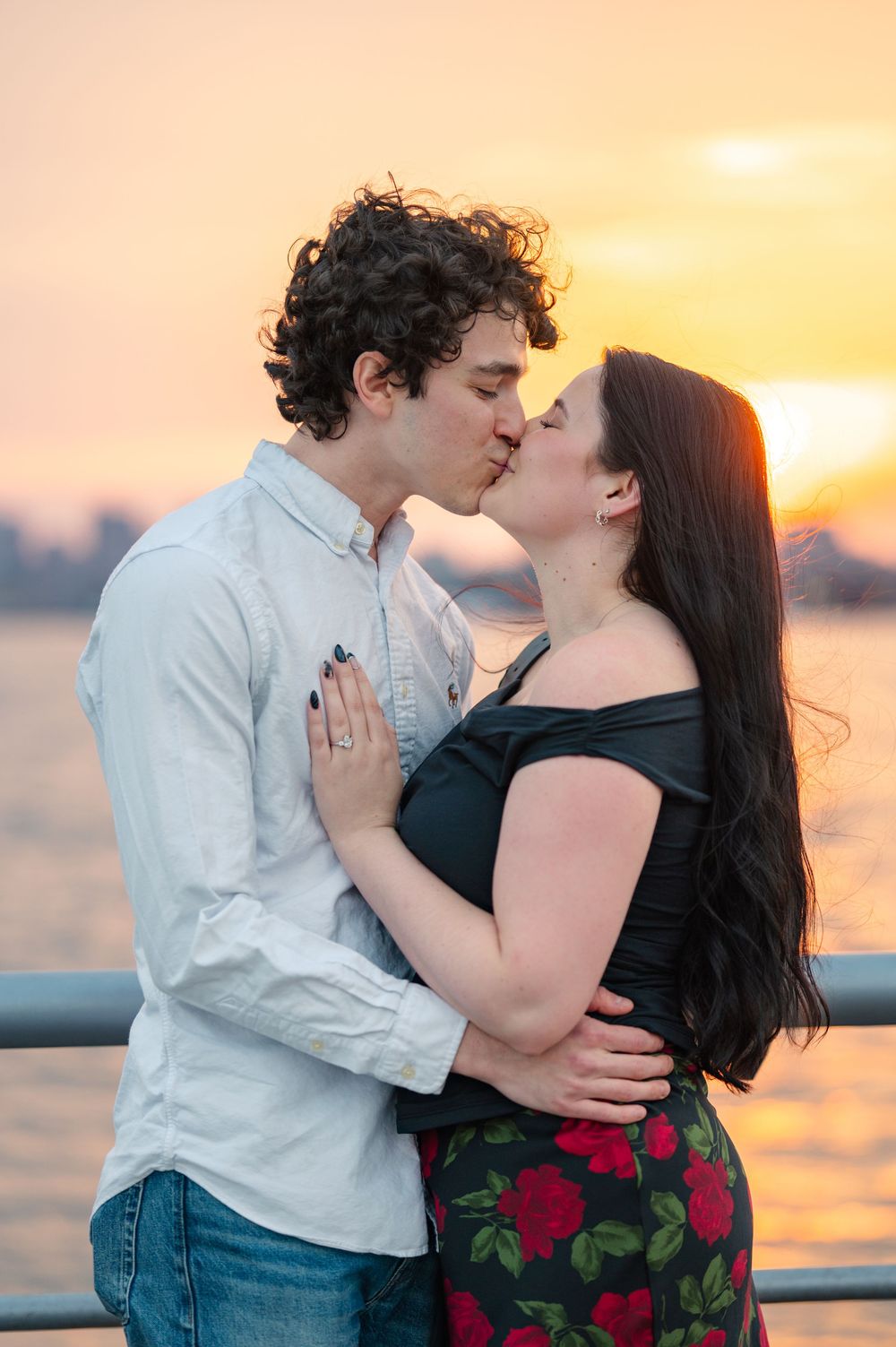 A tender kiss shared between a couple on a pier at sunset with warm golden light reflecting off the water. after their proposal in NYC
