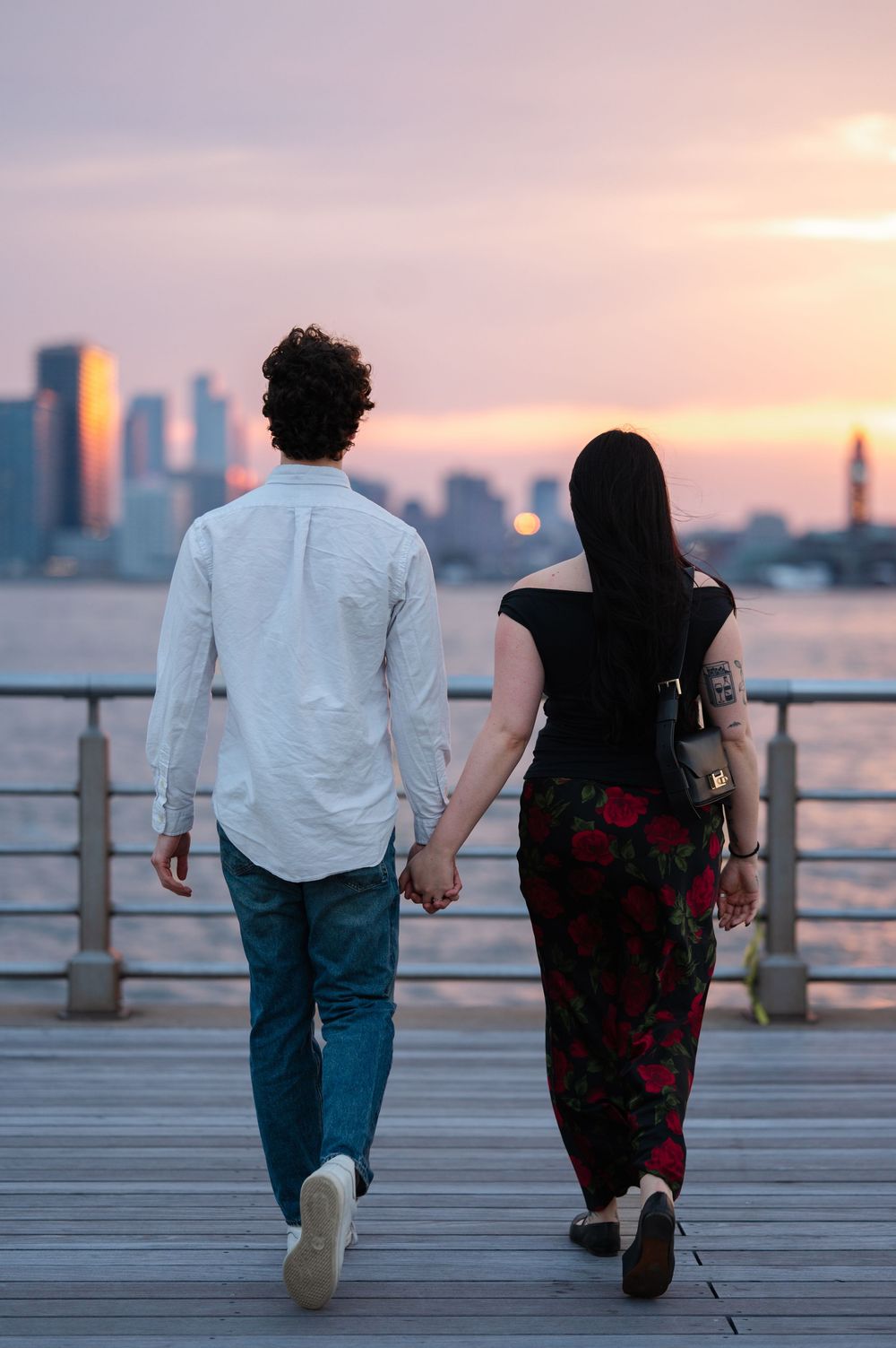 A couple holds hands while walking on a boardwalk pier at sunset with NYC city skyline silhouetted in the background