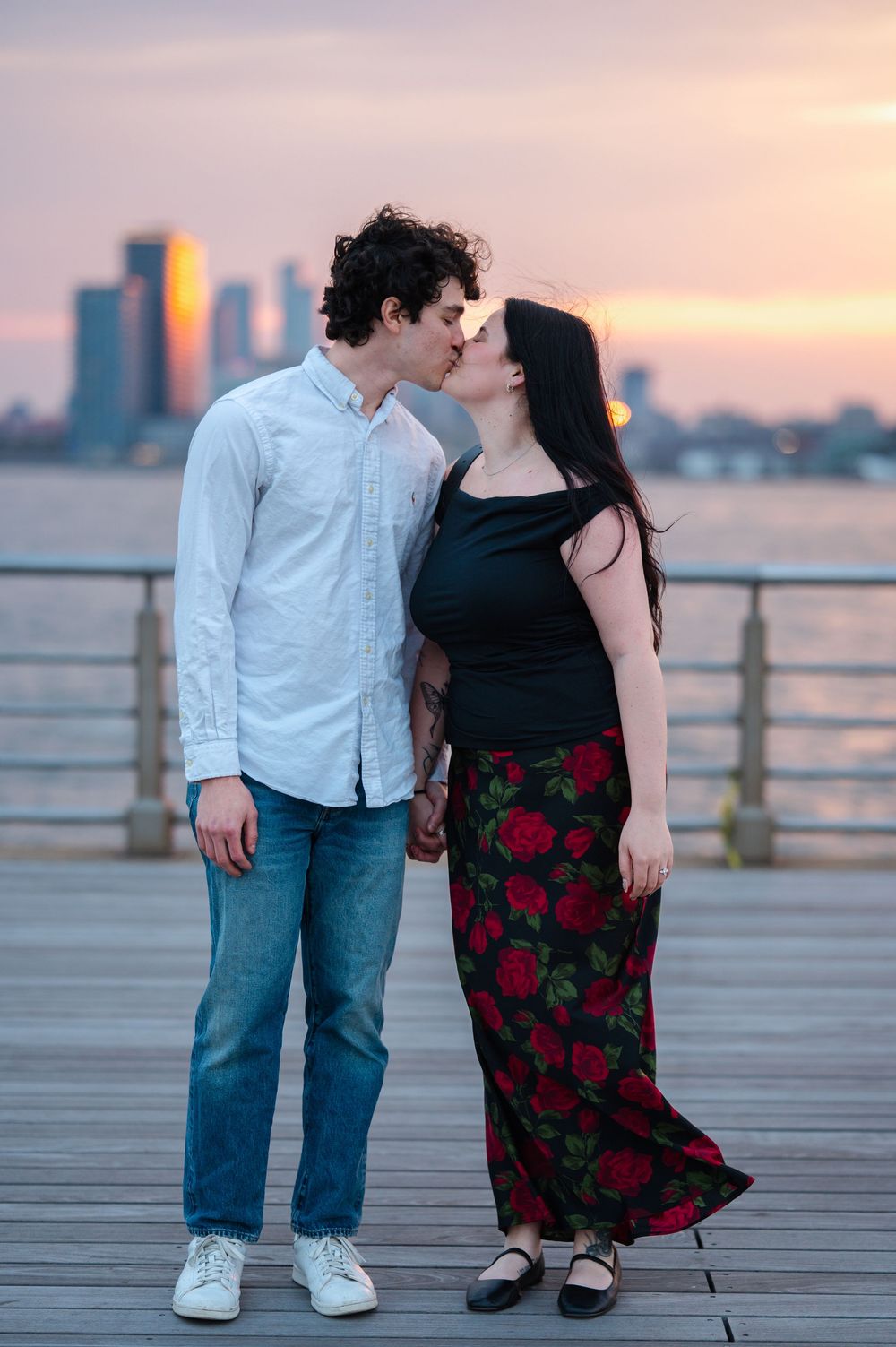 A romantic couple shares a kiss on a boardwalk at sunset with the NYC city skyline in the background during an engagement session