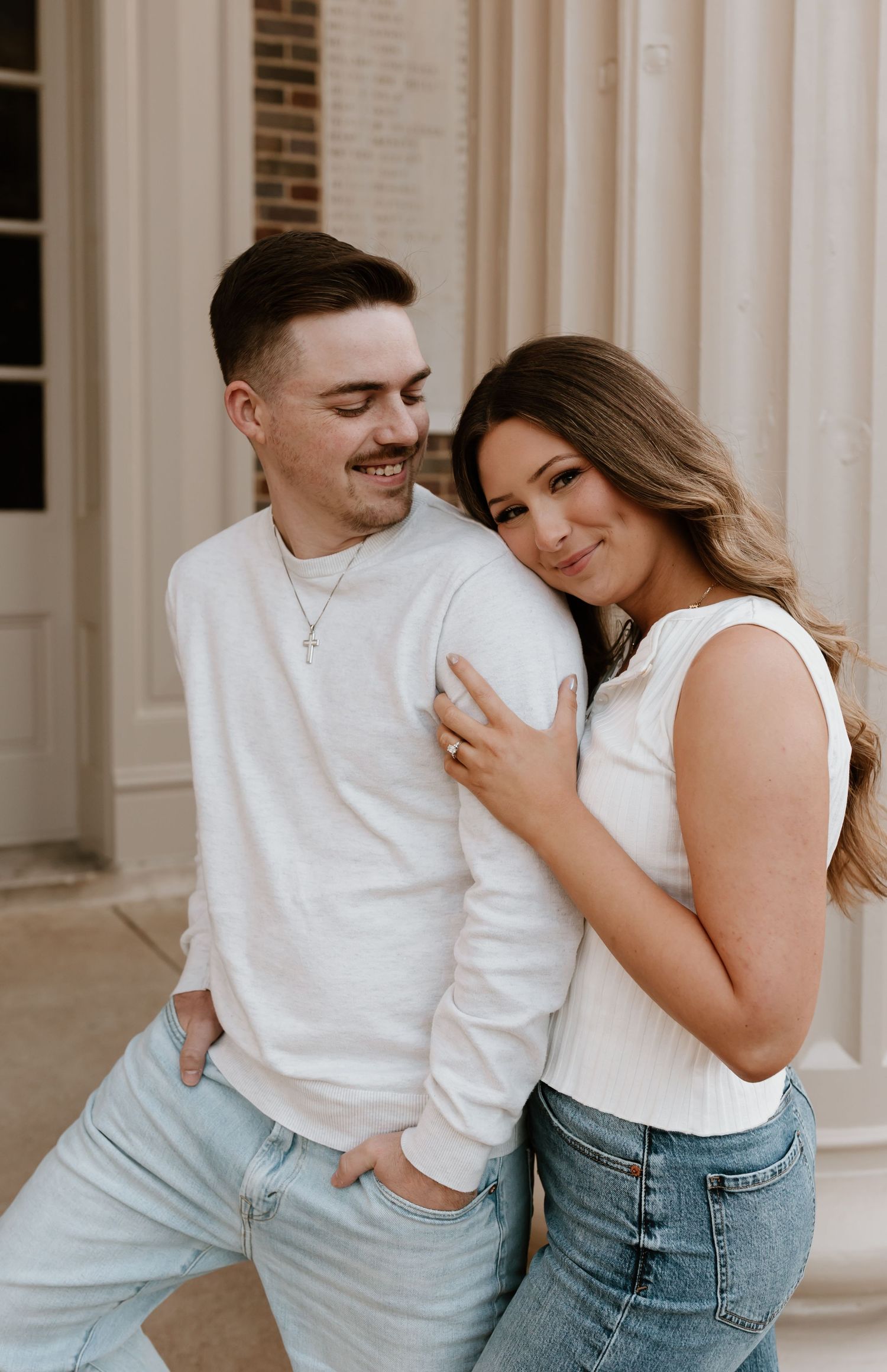 A couple in casual white tops and light denim lean against a brick building while sharing an intimate moment.
