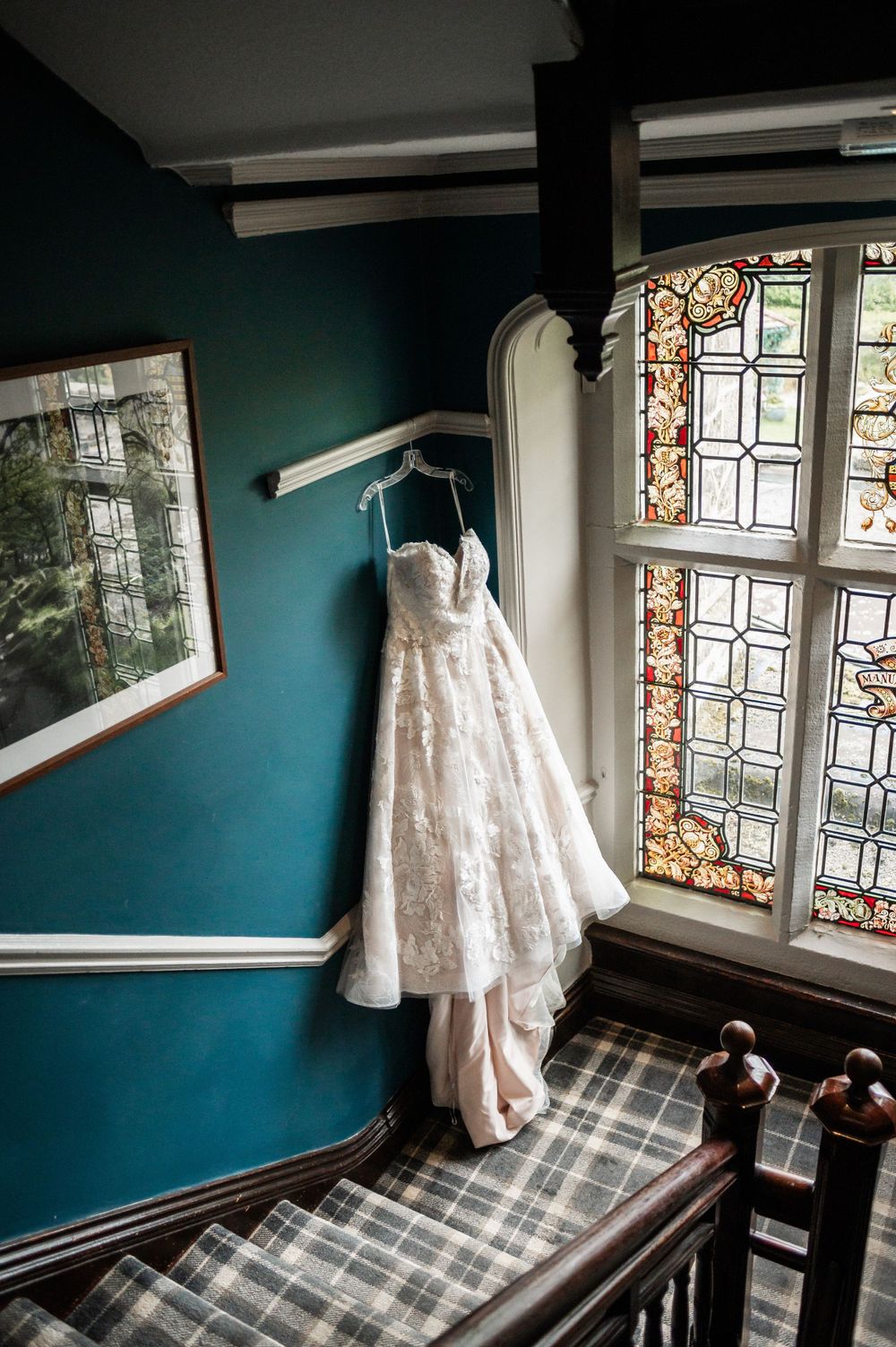A white lace wedding dress hangs on a wall next to a stained glass window in a room with teal walls.