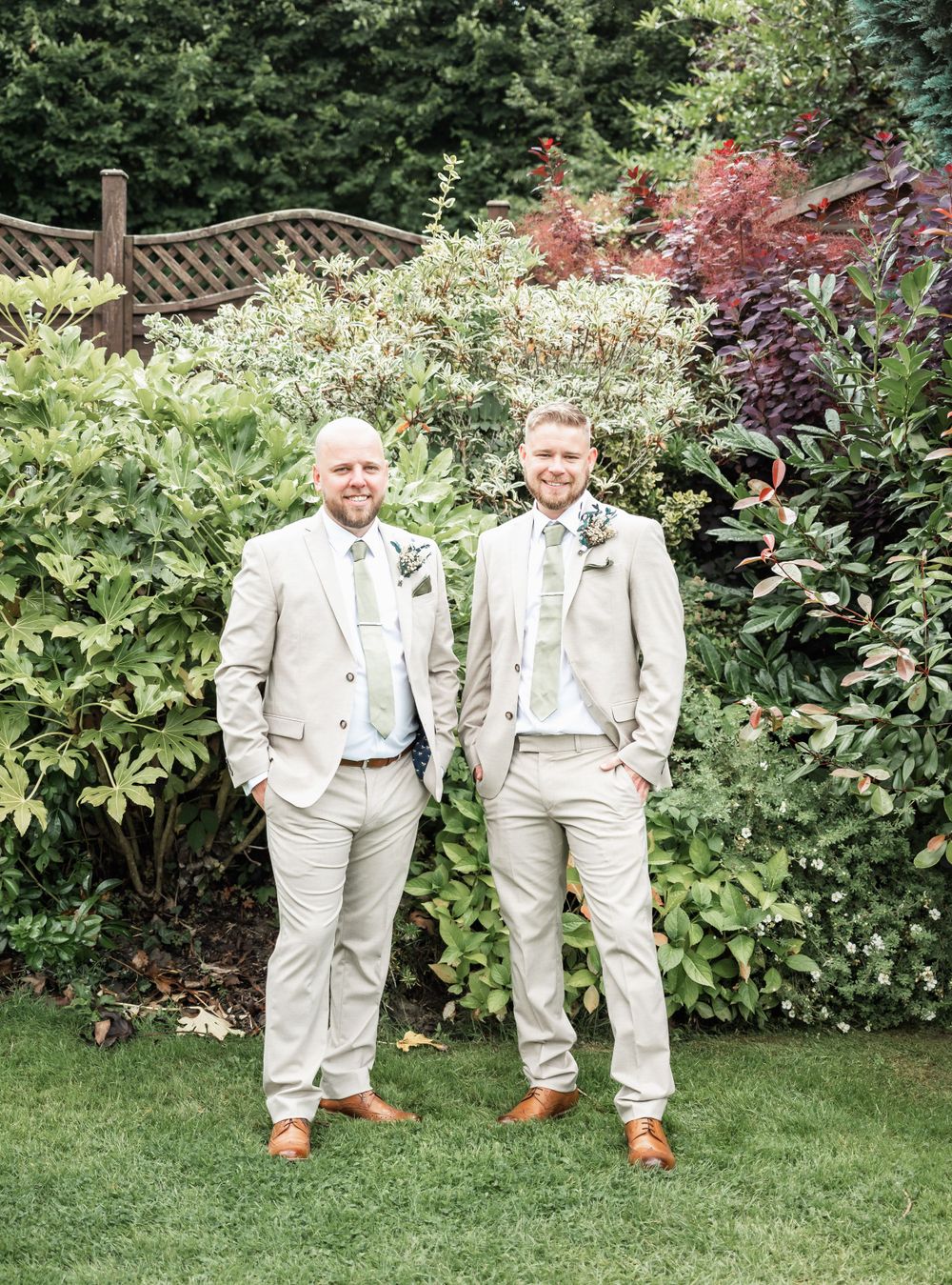 Two groomsmen in light beige suits pose in a lush garden setting.