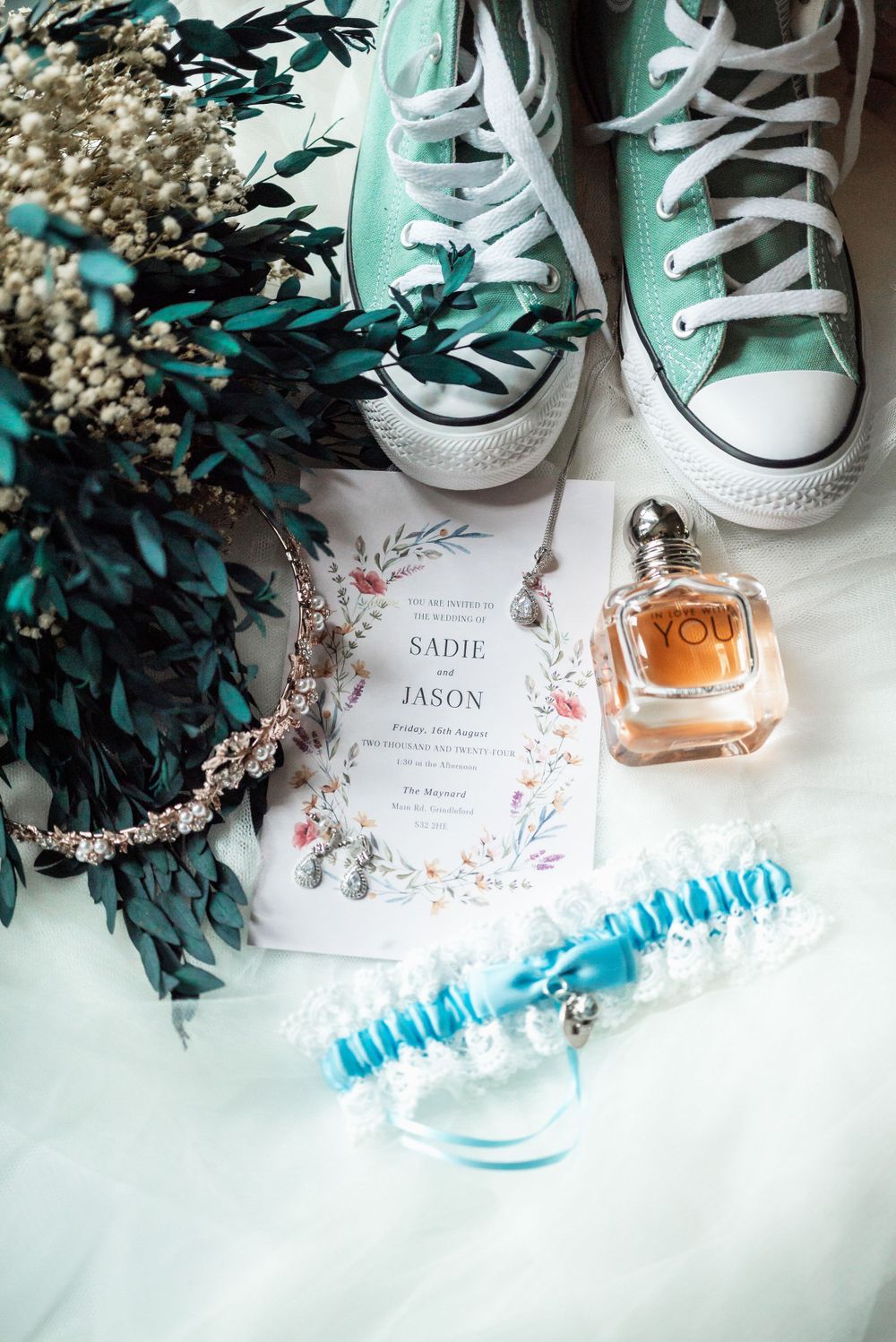 Wedding details including mint sneakers, perfume, invitation and blue garter against a white background.