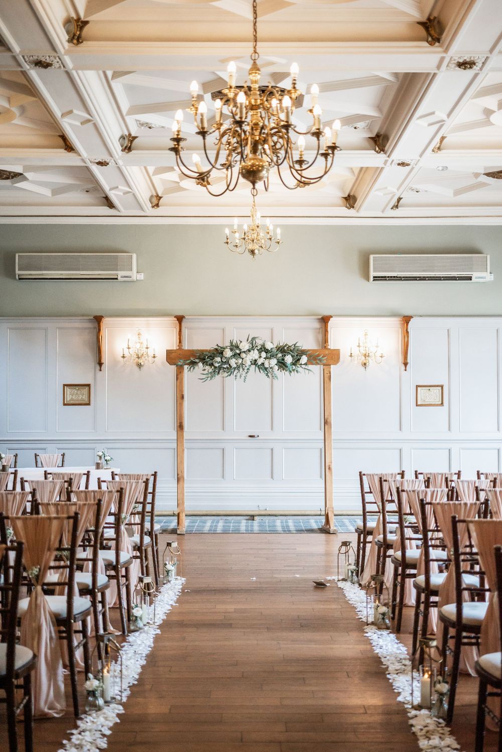 Wedding ceremony setup with wooden chairs and floral arch under elegant chandeliers.