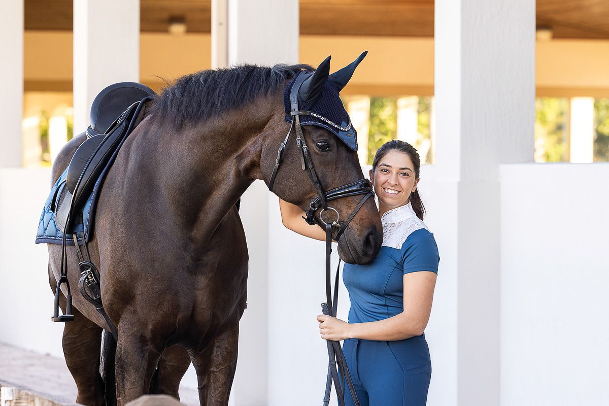 Beautiful dark bay horse with riding tack stands in covered stable area while being groomed.