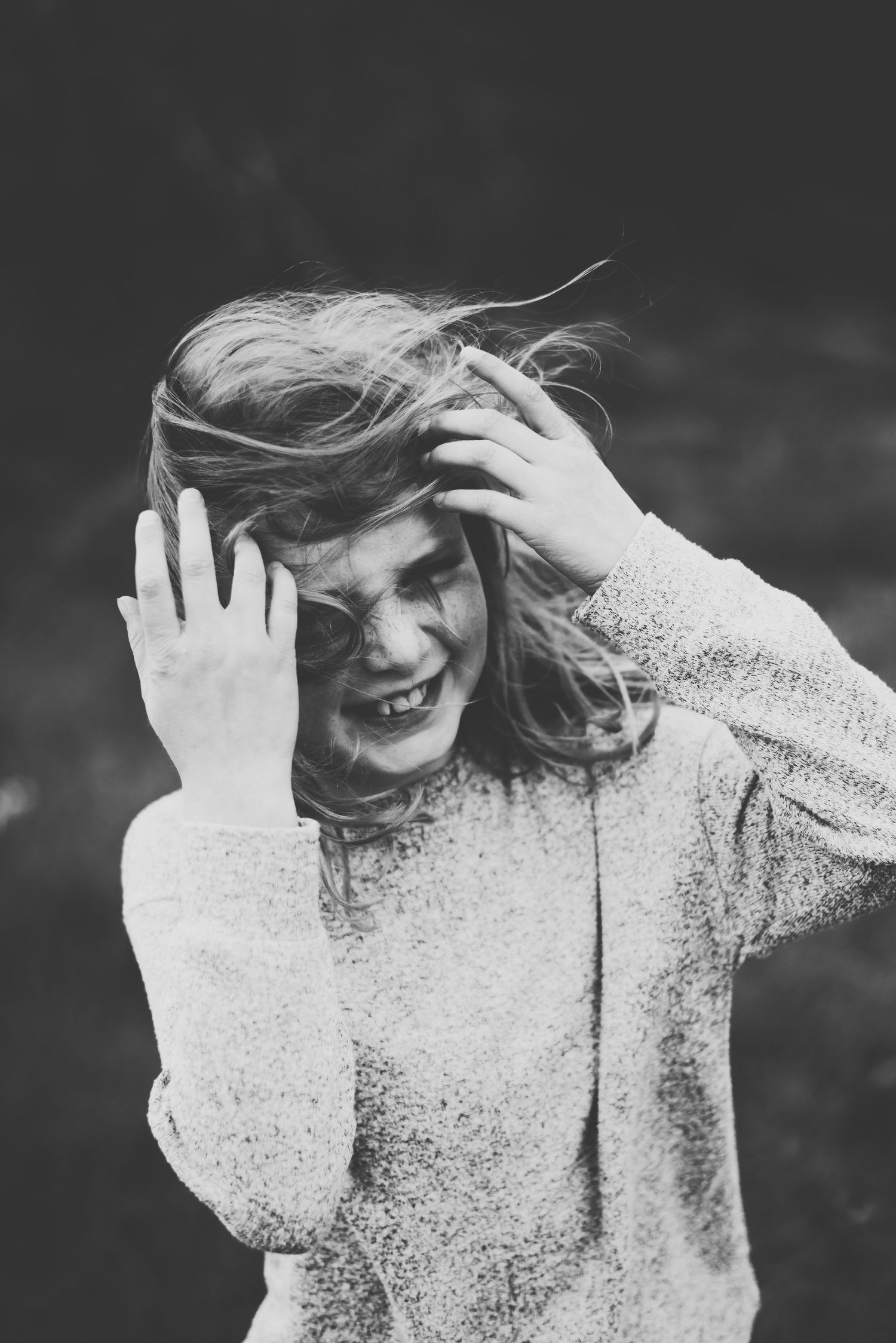 Black and white photo of young girl in sweater laughing with windblown hair while covering face with hands.