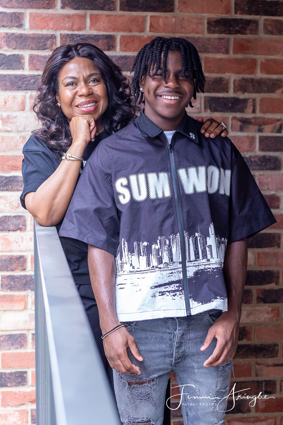 Mother and son in colour co-ordinated clothing pose and smile warmly against a rustic brick wall background.