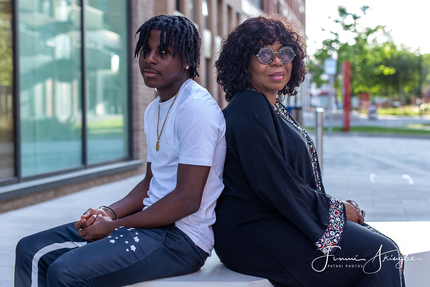 Mother and son sit back-to-back on a bench in an urban setting. Son in a white t-shirt and mum in a black kaftan.