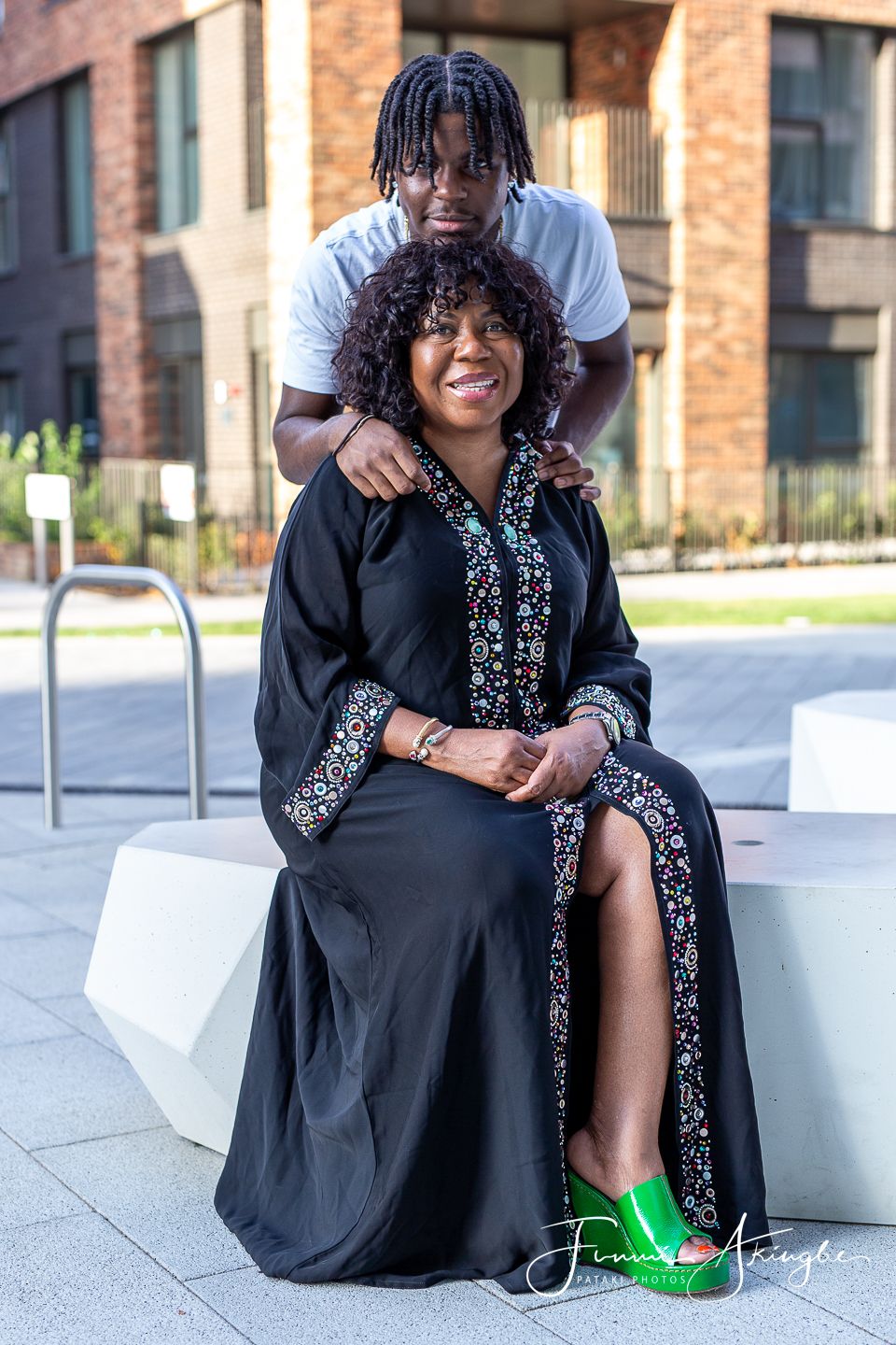 Mother and son portrait. Mum wearing a black embellished kaftan seated on a white bench with her son standing behind.